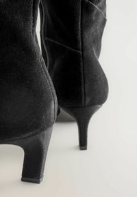 Bottines en daim noir avec un bout pointu et un talon aiguille mince et haut. Fermeture éclair sur le côté et détail de texture douce.