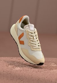 Beige og kremfargede sneakers med oransje detaljer, mesh-overdel, semsket skinn-overlays og en avrundet såle; har snøring og merke-logo på tungen.