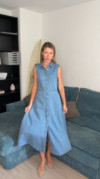 Femme en robe bleue en jean sans manches à boutons, debout dans un salon avec un canapé bleu et une étagère noire derrière elle.