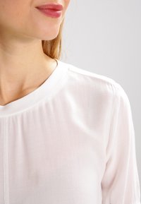 Bílá blůza s kulatým výstřihem, lehkým textura a jemnými skladbami na přední straně. Jednoduchý design bez viditelných vzorů nebo kovových prvků.