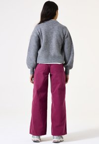 Grau geraffter Pullover mit langen Ärmeln, kombiniert mit locker sitzenden, hoch taillierten rosa Hosen. Modell trägt Sportschuhe.