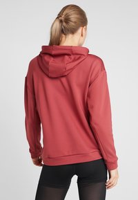 Sweat à capuche rouge avec capuche doublée, tissu doux, épaules tombantes et fentes latérales, associé à un legging noir.