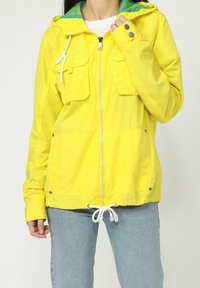 Chaqueta amarilla tipo cortavientos con cremallera en el frontal, dos bolsillos en el pecho, dobladillo ajustable con cordón y forro verde en la capucha interior. Tela ligera.