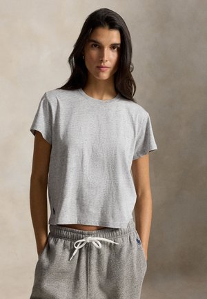 Kvinde med langt mørkt hår iført en lysegrå t-shirt med korte ærmer og grå joggingbukser med hvid snor, hænderne i lommerne.