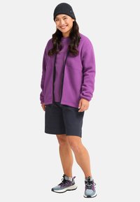 Purple fleece jacket with zipper and front pockets, paired with navy shorts and grey hiking shoes with pink accents. Worn with a black beanie.