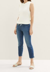 Mujer que lleva una blusa sin mangas de punto blanca, jeans cortos azules con cintura ajustable y sandalias planas beige de tiras, de pie sobre un fondo liso.