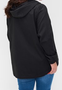 Personne portant une veste à capuche noire avec des poignets élastiques et un jean bleu, vue de dos sur un fond clair uni.