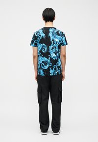 Άτομο που στέκεται με την πλάτη γυρισμένη, φορώντας μαύρο μπλουζάκι με μπλε κυματιστά σχέδια, μαύρο παντελόνι cargo και λευκά αθλητικά παπούτσια σε απλό φόντο.