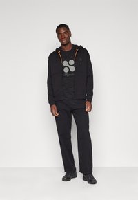 Svart zip-up hoodie med oranga detaljer, svart grafisk t-shirt och svarta byxor. Bäraren står med händerna avslappnat vid sidorna. Svarta skor.