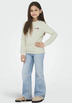 Lichtgroen shirt met lange mouwen en paarse grafisch ontwerp; gecombineerd met wijde blauwe jeans; tan schoenen met rafelige randen. Model staat met de hand op de heup.