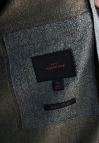 Šedý textilný interiér s čiernym štítkom "NEXT SIGNATURE" a oranžovým textom. Štítok uvádza "VYROBENÉ VO VIETNAMU" a "Talianska tkanina."