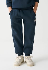 Tamsiai mėlynos sportinės kelnės iš minkštos medžiagos su elastine juosta, šoninėmis kišenėmis ir rezginiu; derinamos su šviesios spalvos sportbačiais.