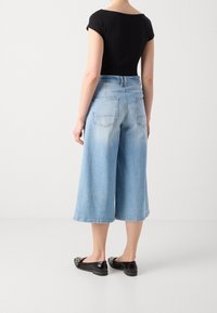 Osoba v bleděmodrých širokých zkrácených džínách, černém krátkém topu a černých slip-on botách, stojící na jednobarevném bílém pozadí.