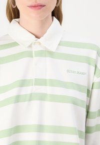 Bílá polo tričko se zelenými vodorovnými pruhy, s límečkem na knoflíky a vyšitým textem "GUESS JEANS" na levé straně.
