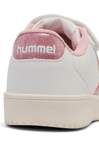 Vit sneaker med en glittrande rosa klack och Hummel-logotyp. Den synliga texturerade sulan och rosa snörning framhäver designen.