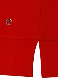 Roter Stoff mit einem kleinen silbernen runden Logo, das ein Kleinbuchstaben "f" und einen vertikalen Schlitz in der Nähe der unteren Kante zeigt.