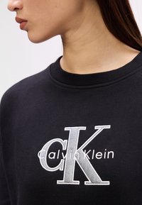 Čierna mikina s vyšitým logom "Calvin Klein" v sivej a bielej farbe, okrúhly výstrih, textúrovaná tkanina, priliehavý strih.