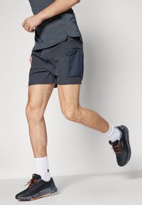 Mörkblå atletiska shorts med en sidficka, kombinerade med svarta löparskor med orange detaljer och vita ankelsockar.
