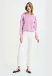 Felpa rosa chiaro realizzata in tessuto morbido, con maniche lunghe, collo rotondo e dettagli testurizzati. Indossata con jeans bianchi e ballerine nere.