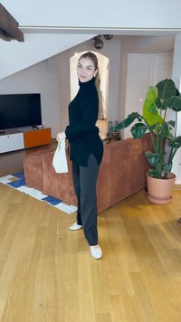 Black ribbed turtleneck sweater, black pants, and white flats. Holding a cream bag. Background includes a brown couch and indoor plants.