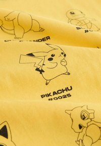 Keltainen kangas, jossa on mustia kuvituksia Pokémon-hahmoista, mukaan lukien Pikachu, Charizard ja Squirtle, sekä tulostetut tunnisteet.