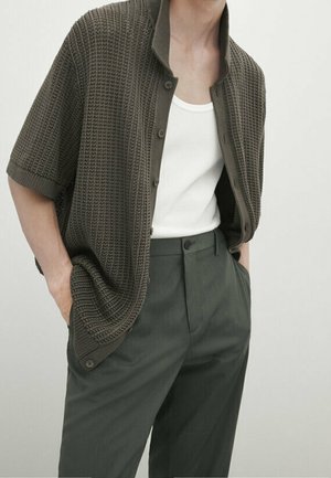 Homme portant un cardigan boutonné à manches courtes texturé vert foncé par-dessus un débardeur blanc et un pantalon taille haute vert foncé, les mains dans les poches.