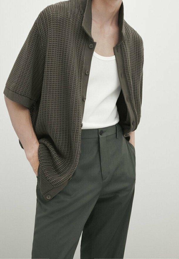 Homme portant un cardigan boutonné à manches courtes texturé vert foncé par-dessus un débardeur blanc et un pantalon taille haute vert foncé, les mains dans les poches.