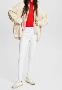 Cazadora bomber crema sobre un cuello de tortuga rojo, combinada con unos jeans ajustados blancos y zapatillas ligeras con un acento crema y amarillo.