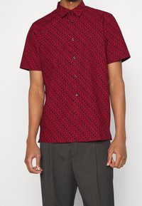 Camisa roja de manga corta con un patrón abstracto negro, cuello abotonado y botones negros, combinada con pantalones grises.