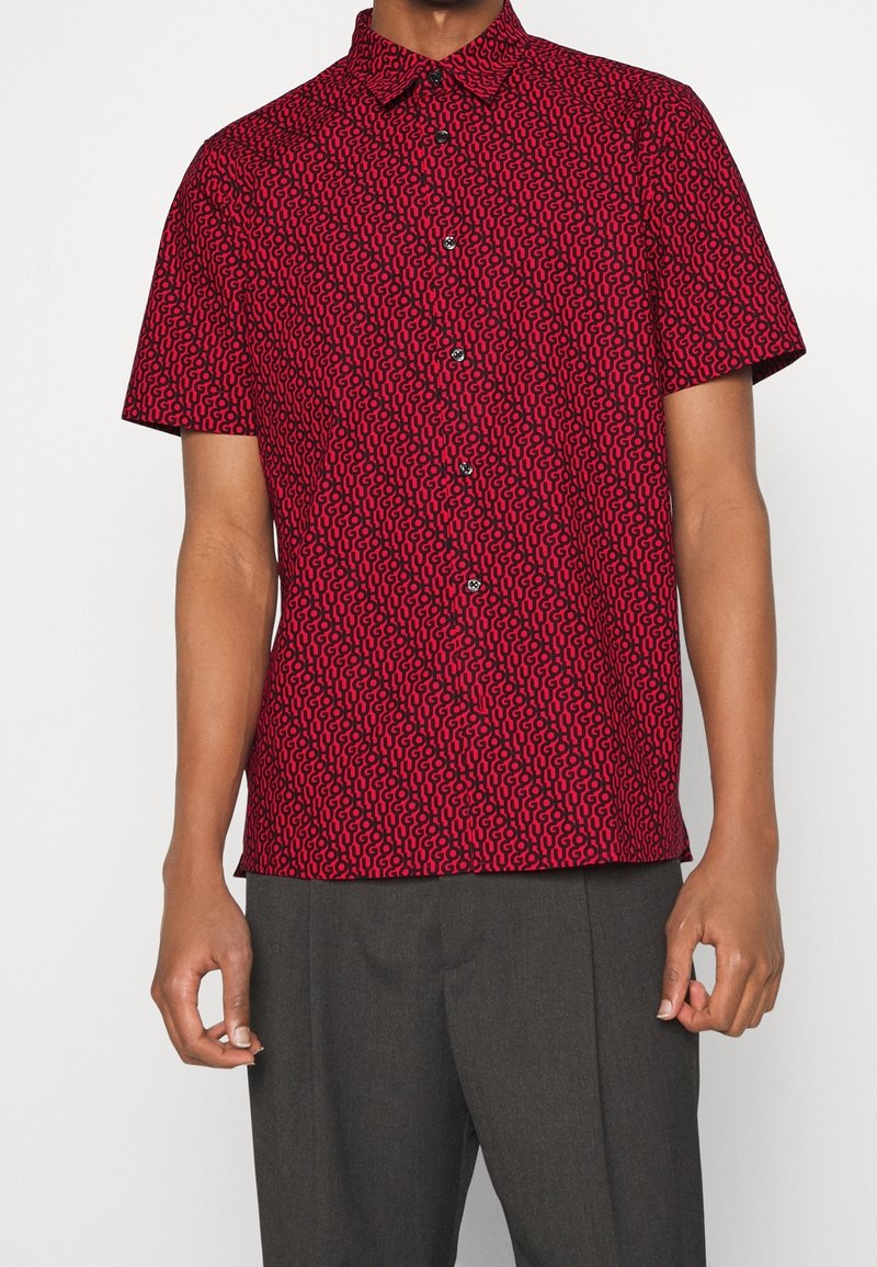 Camisa roja de manga corta con un patrón abstracto negro, cuello abotonado y botones negros, combinada con pantalones grises.