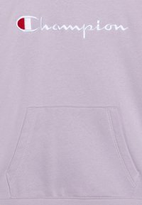 Sudadera lila con un gran logo bordado de "Champion" en rojo y blanco. Incluye un bolsillo frontal tipo canguro. Textura suave.