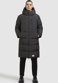 Sort puffjakke med hætte, lang model, lynlåslukning foran og en brystlomme; har tykk polstring og et logobadge på kanten.