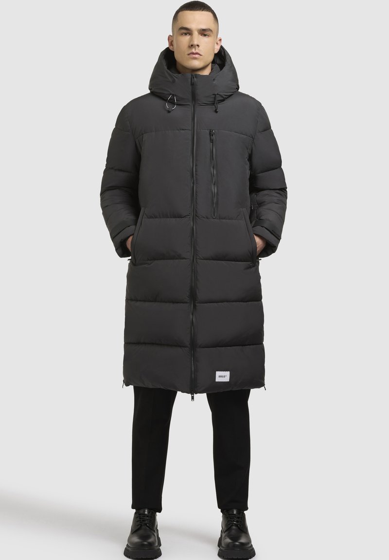 Sort puffjakke med hætte, lang model, lynlåslukning foran og en brystlomme; har tykk polstring og et logobadge på kanten.