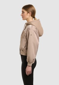 Jeune femme avec une queue de cheval portant une veste à capuche beige et un pantalon noir, debout de profil sur un fond clair.