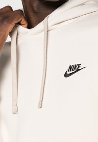 Beige hoodtröja med mjuk textur, med en svart Nike-logotyp på framsidan. Inkluderar dragsko och en känguruficka.