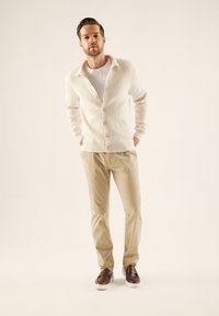 Cremefarbener Cardigan mit strukturiertem Strickmuster und Holzknöpfen, kombiniert mit beigen Hosen und braunen Slippern vor neutralem Hintergrund.