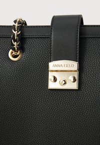 Gros plan d'un sac à main noir texturé avec une bandoulière en chaîne tressée noire et dorée et un fermoir doré gravé "Anna Field".