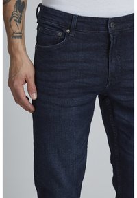 Tamno plave traperice slim fit kroja. Imaju džepove, srebrne detalje i suptilno izbljeđenje. Glatka tekstura s blagim rastezanjem.