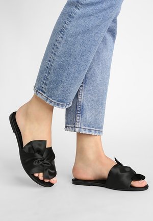 Fødder iført lyseblå kortere jeans og sorte slip-on sandaler med stor satin sløjfe på toppen, stående på en lysegrå gulv.