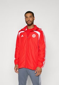 Muž oblečený v červenej tréningovej bunde FC Bayern Mníchov značky Adidas s bielymi pruhmi a sivými nohavicami, stojaci pred jednoduchým svetlým pozadím.