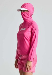 Rosa Sportbekleidung bestehend aus einem Langarmoberteil mit hohem Kragen und angebundenem Mundschutz, kombiniert mit gemusterten Shorts. Inklusive einer weißen Kappe.