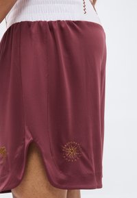 Κοντινό πλάνο βυσσινί αθλητικών σορτς με πλευρικό άνοιγμα και χρυσό κεντημένο λογότυπο πάνω από το τελείωμα στο μηρό ενός ατόμου.