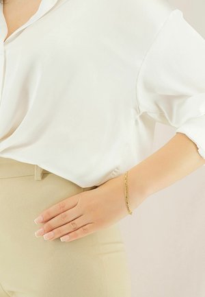 Blusa di seta bianca, pantaloni beige a vita alta e un delicato braccialetto d'oro su una mano appoggiata sui fianchi. Texture lisce e levigate.