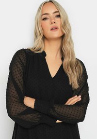 Blusa negra transparente con textura de puntos, escote en V y mangas amplias. Cuenta con puños fruncidos para un ajuste relajado.