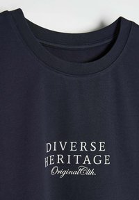 Granatowa bawełniana koszulka z okrągłym dekoltem. Posiada biały nadruk z napisem "DIVERSE HERITAGE OriginalEt h." Miękka faktura, bez wzorów.