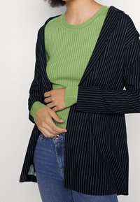 Czarny i biały paskowany blezer nałożony na zielony sweter z prążkami, z długimi rękawami i luźnym krojem, w połączeniu z niebieskimi dżinsami.