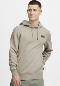 Beige hoodie met een voorzak, verstelbare treksluiting capuchon en een geborduurd logo op de borst. Katoenmix stof met geribde manchetten.