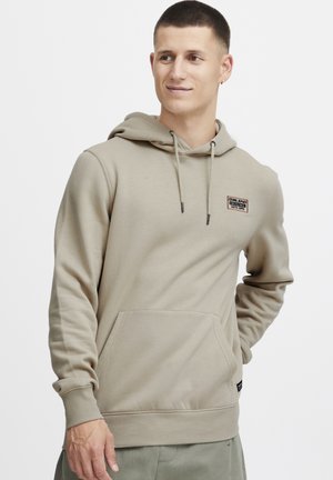 Beige hoodie with a front pocket, adjustable drawstring hood, and a stitched logo on the chest. Cotton blend fabric with ribbed cuffs.
