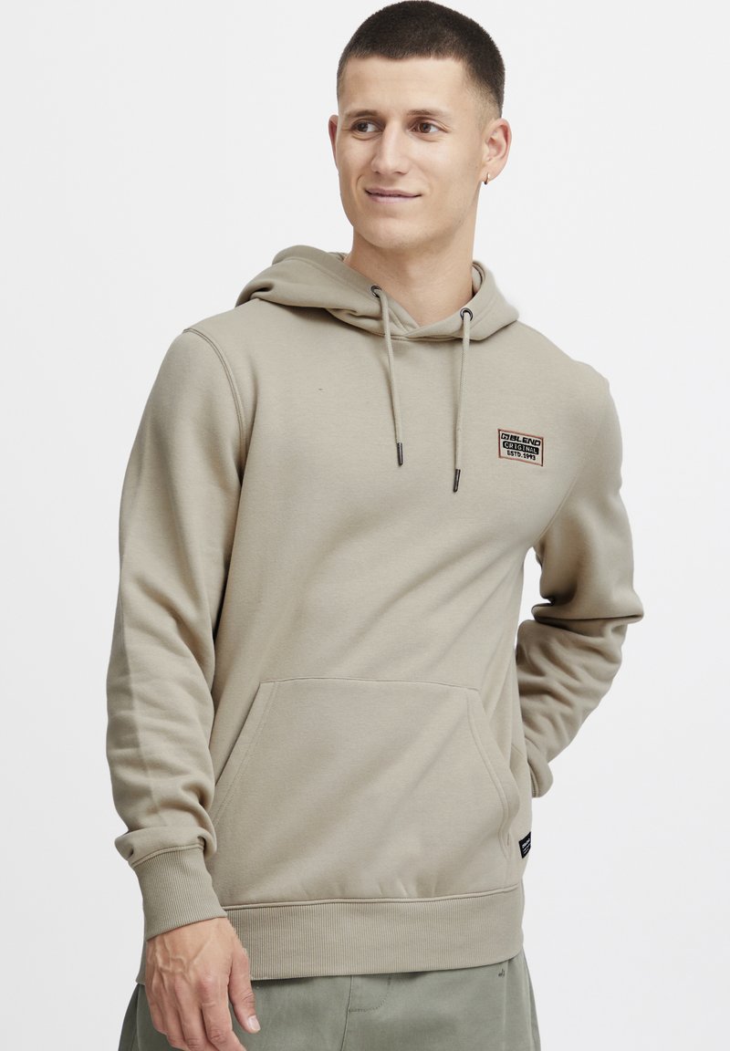 Beige hoodie met een voorzak, verstelbare treksluiting capuchon en een geborduurd logo op de borst. Katoenmix stof met geribde manchetten.
