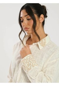 Camisa blanca de tejido texturizado, con un bordado dorado detallado en el collar y los puños. Con botones en la parte delantera y mangas largas.